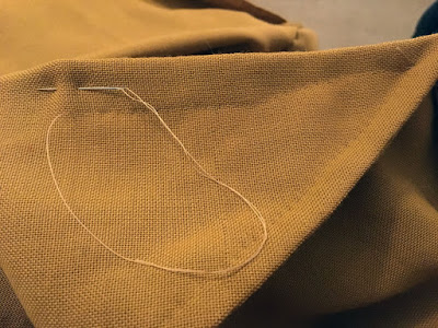 A close-up of a very wide French seam in muddy-gold fabric, with a silver needle threaded with tan thread inserted into the seam allowance at left, ready to continue enclosing the seam.