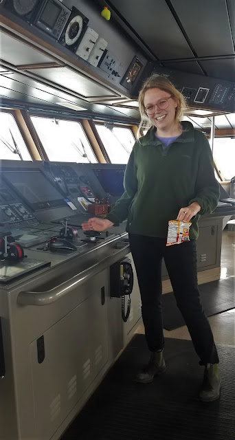 Photo of a smiling young woman standing on the bridge of a ship with her right hand resting on a large, horizontal control panel. Her hand is by a steering wheel about 6 inches in diameter. In the background are windows looking out onto the ocean.