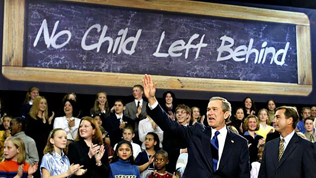 President George W. Bush waves to a crowd. Behind him a sign reads, “No Child Left Behind.”