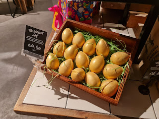 A row of small egg shaped bath bombs coated in gold glitter in a rectangular light brown box next to a black portrait rectangular card that says golden egg bath bomb in white font on a bright background