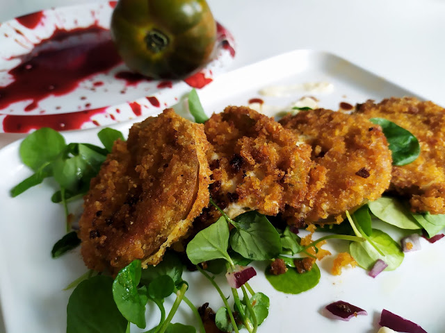 Al fondo un tomate verde sobre un plato que finde estar manchado de sangre. Delante rodajas de  tomate verde empeñado y frito 