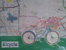 Layered-paper collage with a bus-route map background. Green strip of paper along bottom, roughly one-quarter height of total image. In lower left corner, the word 'Bicycle' has been pasted, in blue letters against white background. The cut-out image of a blue, step-through framed bicycle, shown from side view with multi-colored flowers in a handlebar basket, is positioned on the green strip as if it were being ridden over the green 'ground.' Behind it, to the left in picture, is a cut-out paper image of a fabric butterfly. In the upper-right corner is a yellow half-sun. The artist's initials, 'CMP,' are rendered in individual cut-out letters, black type against white background, in the lower-right corner of the picture.