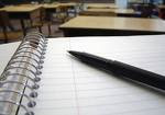 This is a picture of an open notebook on a desk and and a pen laying on the blank page and  other desks in the classroom in the background 