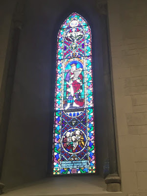 A single, tall gothic arched stained-glass window, with a king in a red mantle holding a harp in the center medallion, and smaller medallions above and below. The border is composed of turquoise, pink, and purple vining elements, with red and purple crossed panels of squares dividing the medallions.