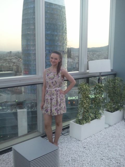 girl with brown ponytail wears cream floral dress with thin white belt against Barcelona skyline