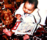 Baby sits up in patterned couch, rumpled magazine across her body as she looks down towards the words.