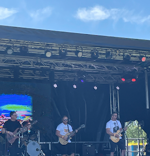 An outdoor stage, a three-piece band, two musicians with guitars, amps and sound stuff all around, and perfect blue skies above.