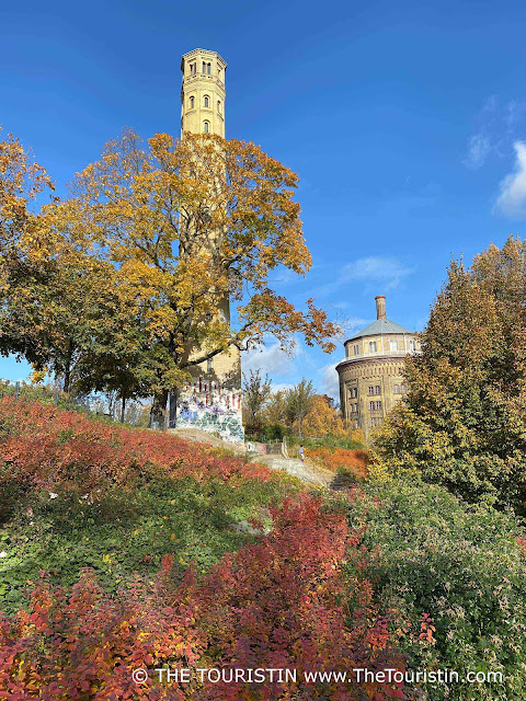 A water tower and a tall tower on a hill covered with trees and shrubs in colourful autumn leaves, under a bright blue sky.