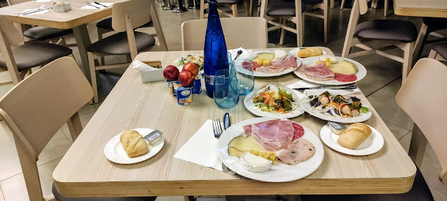 A square table in an empty restaurant, laid with three plates containing cold meats and cheeses, three of sald and another three small plates with a bread roll and butter on.