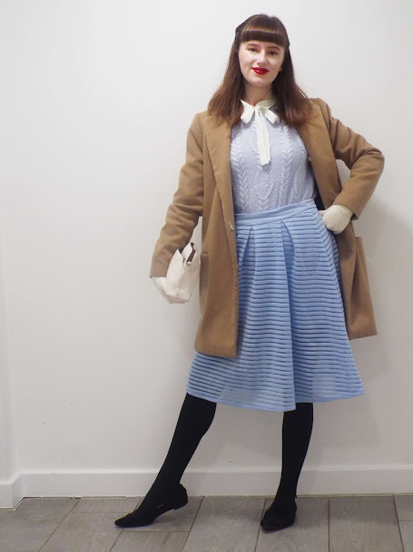 Ellie, poses smiling in 40's styled white pussy bow shirt and baby blue jumper and round midi skirt, paired with dainty white gloves, camel beige smart coat, cream handbag, black tights, pointed toe black slip on shoes and hair styled with 40s twists and a red lip.