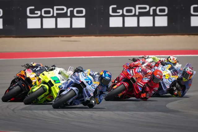Grupo de pilotos de MotoGP, incluyendo a Marc y Alex Márquez, inclinados en una curva durante una intensa batalla.