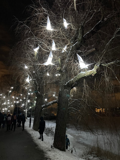 Sculptural origami birds are suspended in an avenue of trees. They are lit up and glow a bright white, as people in thick coats and hats walk through a snowy park.