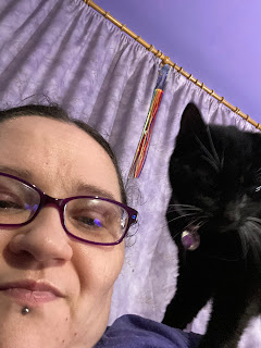 A close up selfie taken by a brunette, bespectacled, middle aged white woman with a black cat with white whiskers standing on her left shoulder like a parrot. They're both looking at the camera; and the woman looks absolutely exhausted.