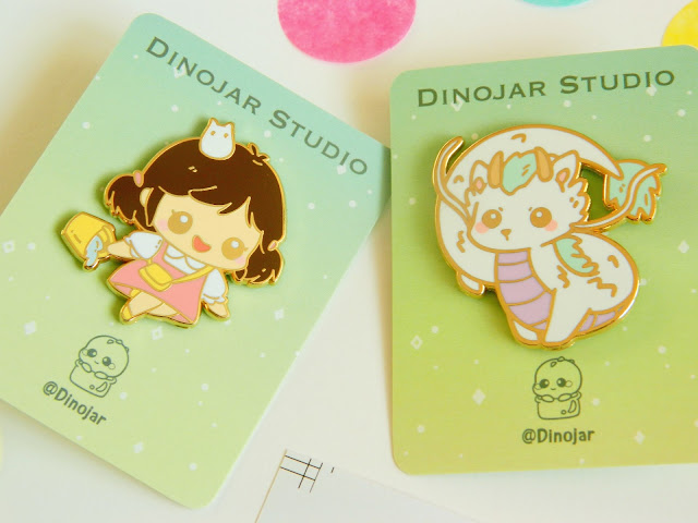 A photo showing two enamel pins by Dinojar. They are of the Studio Ghibli characters, Mei and Dragon Haku. They are backed on gold enamel and are super cute!