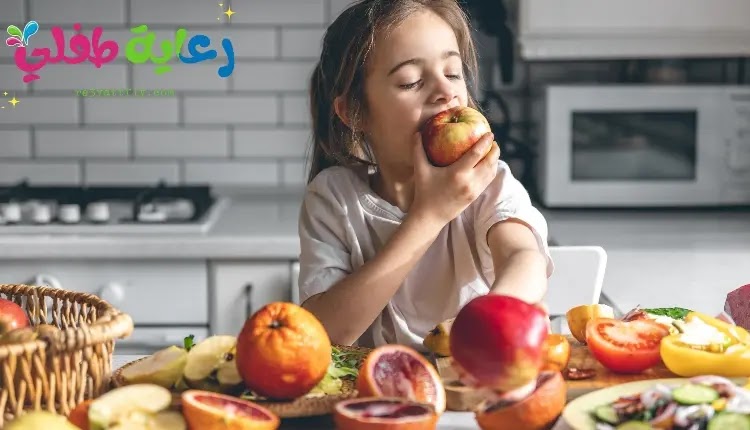 صورة فتاة تأكل من تفاحة، وأمامها منضدة عليها مجموعة متنوعة من الفواكه، تعبيرا عن فوائد الفواكه للأطفال.