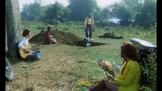 a group on hippie bikers sit around as their dead friend sits in his grave