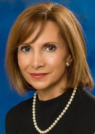 Portrait of Antonia Gavrihel set against a professional blue backdrop. She is looking at the camera, smiling slightly, in a black sweater with a string of pearls around her neck.