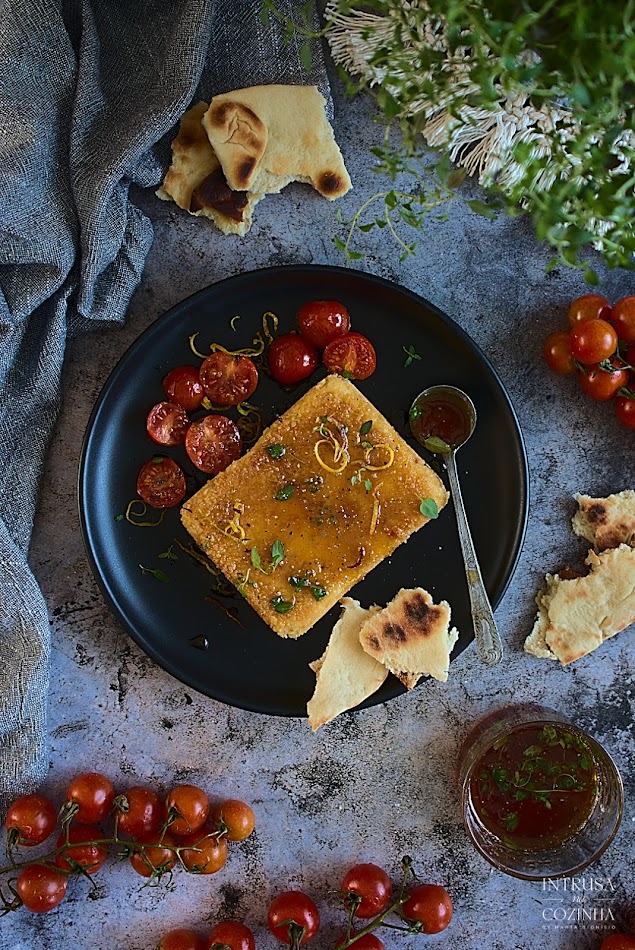 Queijo panado num prato preto com tomates cherries à volta, com titas de limão e folhas de tomilho