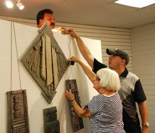 MCC student Dave Hatch, behind wall, helps artist Janet McIntyre and exhibit designer Greg Heil install a series of hanging sculptures in her show 'Wall Jewelry.'