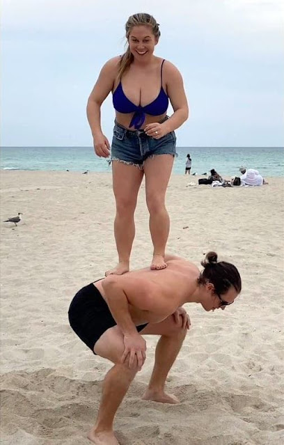 a small woman standing on the back of a crouching big man on the beach.