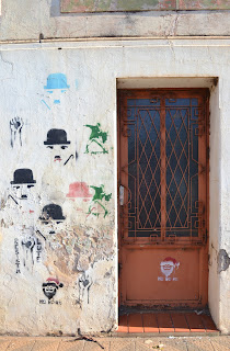 À direita há uma porta marrom de uma casa abandonada. À esquerda há vários desenhos na parede: cinco reproduzem o personagem Charlie Chaplin, um o Papai Noel com a escrita “Ho ho ho” abaixo, três pinturas de braços levantados e as palavras “lute” e “resista”. As intervenções urbanas utilizadas nesta parede são chamadas de estêncil. A técnica necessita da utilização de pranchas com desenhos vazados que é por onde passa a tinta ou o spray. Esta casa se encontra na Avenida Presidente Vargas, no centro da cidade, próxima ao Hospital Ivan Goulart