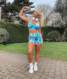 A young girl wearing headphones around her neck and a matching blue and white gym outfit- shorts and top. She is flexing her bicep and smiling.
