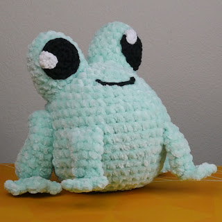 A crochet frog plushie made with fluffy yarn in mint green facing the camera.