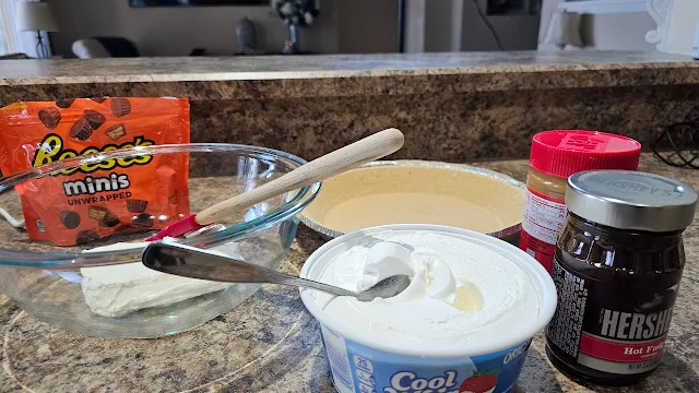 Reese's peanut butter cups, a jar of peanut butter, cream cheese in a bowl, hot fudge sauce, Cool Whip ingredients on a counter.