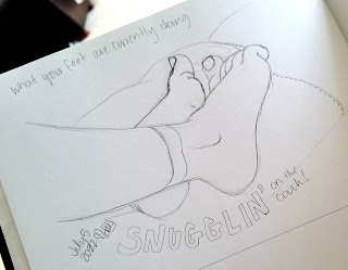A photograph of a pencil drawing in a sketchbook. The drawing is of two feet, resting together on a couch. The words under the feet say "Snugglin' on the couch."
