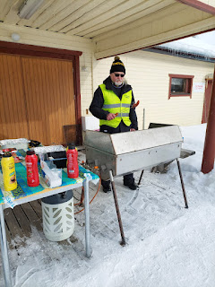 Kuvassa mieshenkilö grillin ääressä makkaranpaistopuuhissa, katoksen alla. Taustalla puinen rakennus.