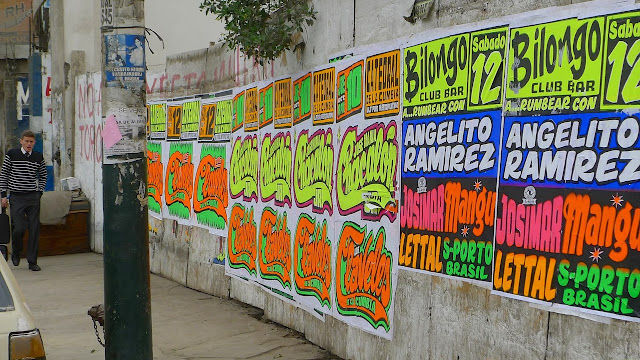 Muro cubierto de afiches con "estética chicha": colores flúo, letras grandes y estilo psicodélico años 70, en una calle de Lima. Se ve a un transeúnte pasando por la vereda.