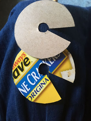 Two circles of cheap saltine cracker box, each with a tiny circle cut from the center and a small wedge flaring from the inner circle to the outer edge. The top piece shows plain cardboard, while the bottom shows the bright yellow and blue package design of the cracker box.