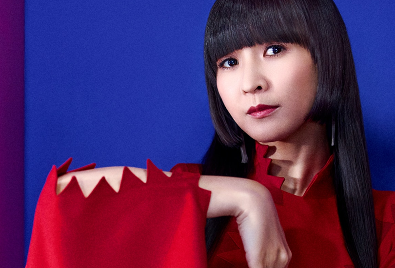 Kashiyuka of Perfume, posing against a blue backdrop, wearing a red dress.