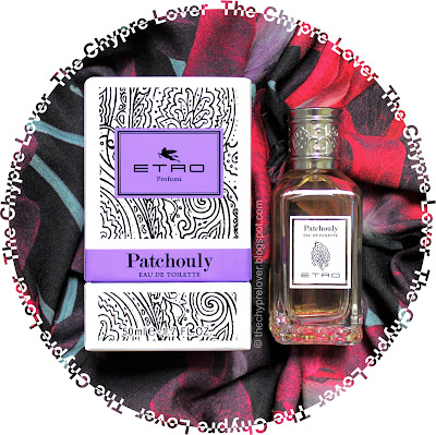 The bottle and the paisley-laden box (black lines on a white background) lying flat on a multicoloured blouse (red, magenta and black)