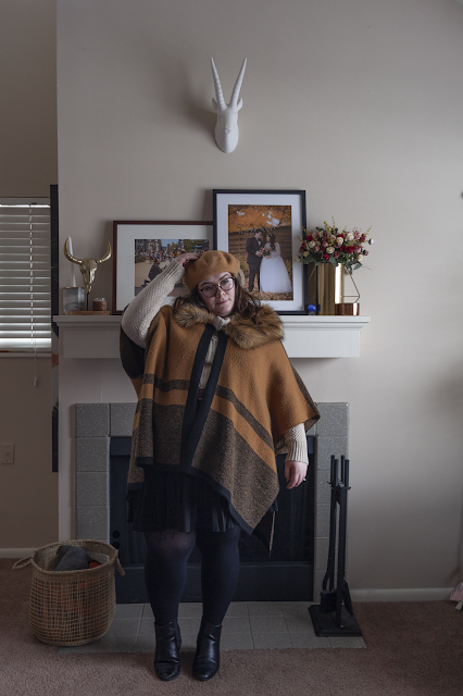 An outfit consisting of a tan brown beret, brown and lack poncho over a cream cowl neck sweater tucked into a black pleated mini skirt and black ankle boots.