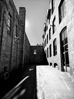 A black and white photo of an alley surrounded by brick walls and half of it is in sunlight and half of it is in shade.