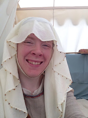 A young white woman in a grey dress and white veil with a beaded edge, inside a white canvas tent.