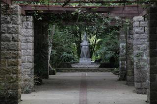 Foto da entrada principal do Orquidário de Santos. Ao ar livre, um amplo corredor central pavimentado com grandes placas de concreto, lateralizado por colunas de pedras. Da base de cada coluna, uma planta trepadeira sobe e envolve o fechamento superior, composto por um pergolado ripado. Ao fundo, um pouco a frente da densa vegetação a escultura de granito, em estilo art decó,  “Ninfa de Náiade”ou “Mulher de Pedra”, de frente, sentada e desnuda sobre uma base, apoia os cotovelos sobre os joelhos unidos e as pernas em X, a ninfa de Náiade eleva uma ânfora de onde verte a água que a banha.  Curiosidade: A obra é do escultor João Batista Ferri.