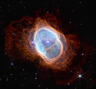 NGC 3132 - The Southern Ring Nebula - NIRCam Image - A planetary nebula, seen by the Webb telescope’s NIRCam instrument, against the blackness of space, with points of starlight behind it. The nebula itself is shaped like an irregular oval, with lacy, reddish orange plumes of gas and dust. Further inside the circle, the gas and dust glows bright blue. A glowing white ring separates the red and blue gases. In the center of the rings are two stars, one glowing much brighter than the other, with diffraction spikes radiating out from it.