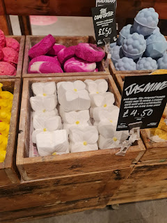 A zoomed in photo of some white flower square shaped bath bombs in a light brown square box on a light brown table with a black card that says jasmine in white font on a bright background