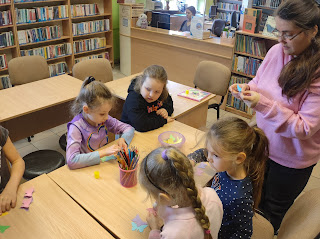 Sala biblioteczna – Pani bibliotekarka stoi przy stoliku, przy którym siedzą dzieci w ręce trzyma papierowego niepieskiego motylka. 4 dziewczynki siedzące przy stoliku wykonują swoje motylki.