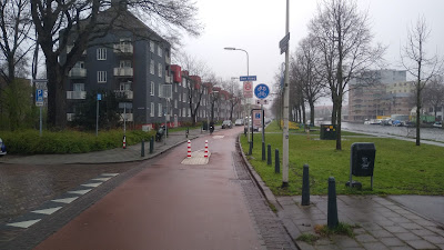 A street with flats to the left and a canal to the right. A red road is blocked to traffic by bollards that people can cycle through.