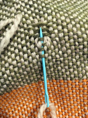 Close up of the same light green and white block, with a marigold and white block showing below. The light blue plastic yarn needle is threaded with white yarn, and inserted under a few green threads below the floats. The point of the needle rests just over the first light green float.