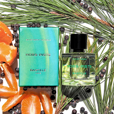 The green-blue box and glass bottle of Hors-Piste by Bastille lying flat on pine neddles next to juniper berries and slices of mandarin orange