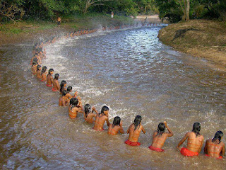 Foto em dia claro. Inumeráveis jovens índios sentados nas águas rasas de um rio margeado por vegetação rasteira e densa, formam um imenso semicírculo que parte do canto inferior direito a perder de vista ao fundo à direita. Todos tem cabelos escuros lisos abaixo dos ombros, no dorso moreno exibem três listas horizontais pintadas em vermelho no mesmo tom das sungas; as mãos agitam as águas que sobem e caem como uma divertida chuva salpicando as águas em branco.