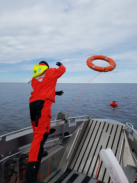 Pelastautumispukuinen henkilö heittää pelastusrengasta meressä kelluvalle henkilölle veneestä