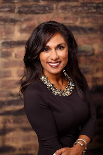 Image of a smiling woman with dark hair wearing a brown blouse and standing in front of a brick wall