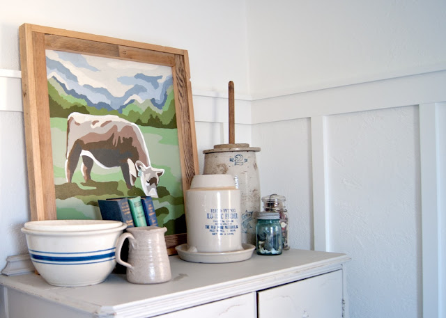 Farmhouse Master Bedroom Reveal - Benjamin Moore Simply White, Paint by number cow painting . antique crocks, red wing, chicken feeder, books, mason jars