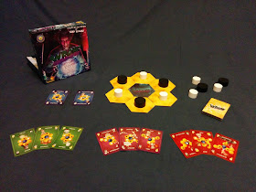 The game components. The box, with its cover art of a stereotypical mad scientist holding a large glowing sphere that contains the schematic image of an atom (the circle representing the nucleus and three oval orbits of the electrons around it), sits next to the tokens, tiles, and cards. In the centre is the main playing area: six yellow hexagonal tokens surrounding a blue hexagonal token with the 'Wrong Chemistry' title on it. The yellow tiles each have a large round wooden token on them, alternating black and white. A few extra tokens sit nearby, with the draw pile of the card deck next to them. On the other side of the tiles are two blue cards, the 'Restartium' and 'Extramovium' cards. In the front are nine cards from the deck, arranged in three groups. The first group has three sample green cards, worth one point each. The next group has three purplish-red cards, each worth two points. The last group of three contains red cards with three points each.