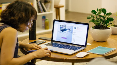 A female marketer working with a computer surrounded with potted plant in a white pot, notepads, mousepad and smart phones.